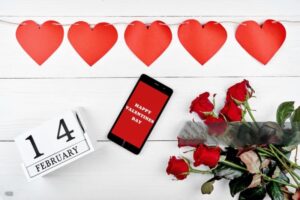 Garland of red hearts, bouquet of roses, cellphone with sign Happy Valentines day and february 14 wooden calendar, copy space. Greeting card mockup. Love concept. Top view, flat lay